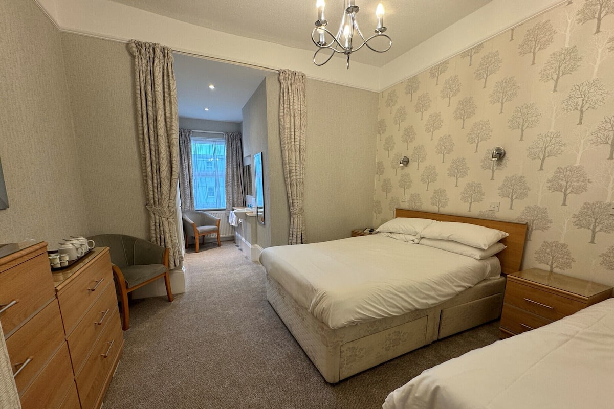 A bedroom with a double bed, a wooden dresser, a chair, a desk, and a chandelier.