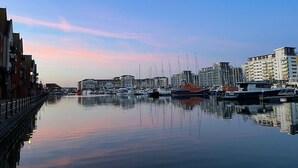 Un port de plaisance avec des bateaux amarrés, des bâtiments le long de la rive et une surface d’eau calme qui reflète le ciel.