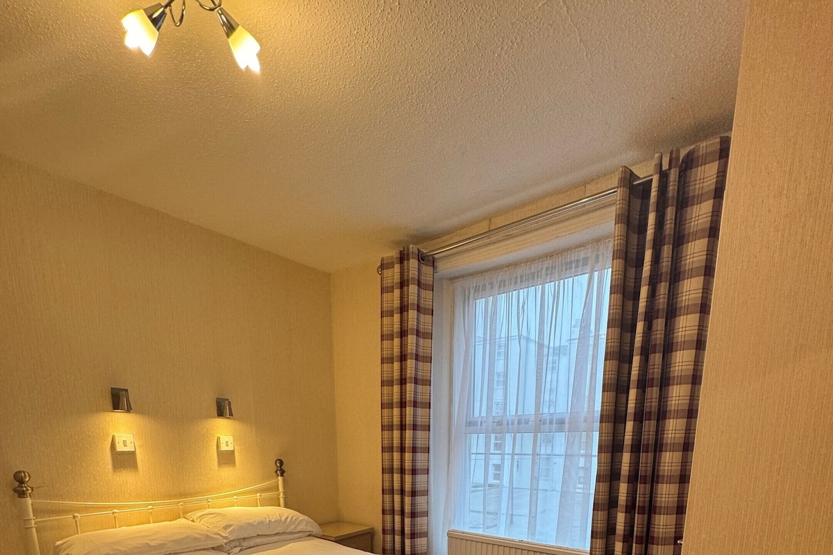 A bedroom with a bed, two bedside lamps, a window with curtains, and a radiator.