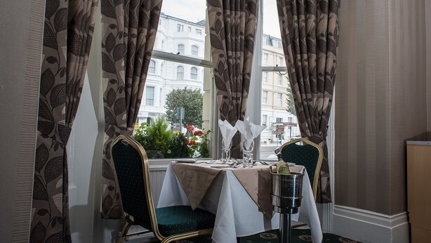 Une table à manger dressée avec une bouteille de champagne, des verres et des couverts, donnant sur des immeubles à travers de grandes fenêtres.