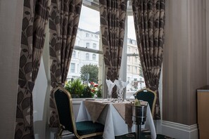 A dining table set with a bottle of champagne, glasses, and cutlery, with a view of buildings through large windows. - Haddon Hall Hotel (Eastbourne)