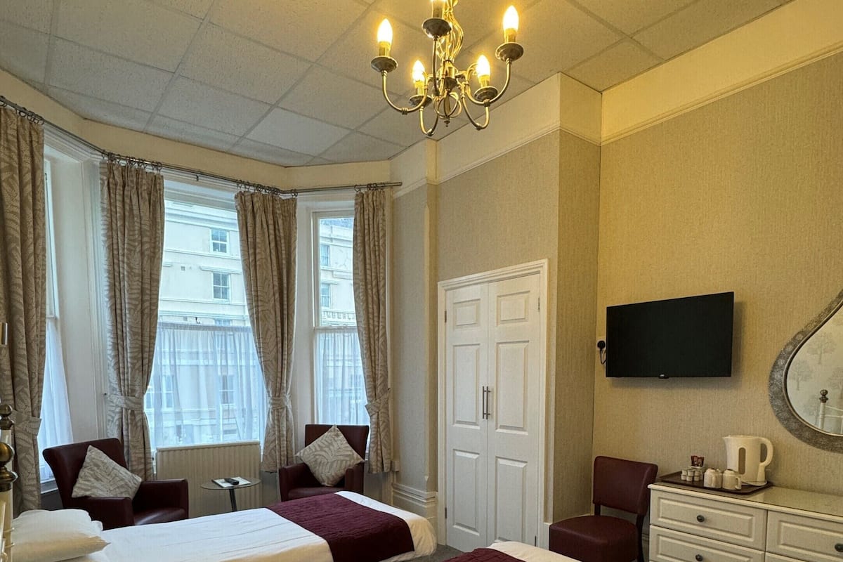 A hotel room with two beds, a chandelier, a television, and a view of buildings through the window.