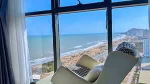 Una stanza con una grande finestra vista spiaggia e oceano. C'è una sedia con un cuscino decorato e un oggetto decorativo sul davanzale.