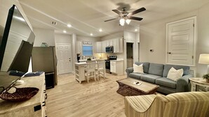 A modern living room with a grey sofa, a coffee table, and a TV. There is a kitchen area with white cabinets and a microwave. - Coastal Dreamer-Family Friendly, 2 Kitchens, Golf Cart Included, Close to Beach (Port St. Joe)