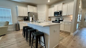 A modern kitchen with white cabinets, a central island with stools, stainless steel appliances, and a window with blinds. - Coastal Dreamer-Family Friendly, 2 Kitchens, Golf Cart Included, Close to Beach (Port St. Joe)
