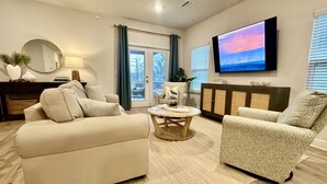 A living room with a beige sofa, patterned armchair, and a coffee table. A TV is mounted on the wall, and there is a cabinet with decorative items. - Coastal Dreamer-Family Friendly, 2 Kitchens, Golf Cart Included, Close to Beach (Port St. Joe)