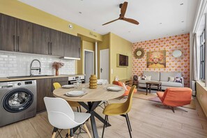 A modern kitchen with a built-in washing machine, a dining area with a round table, and a living space with a sofa and a colorful wall.
