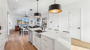 A modern kitchen with white cabinetry, a large island with a sink, pendant lighting, and a view of the outdoors through large windows.