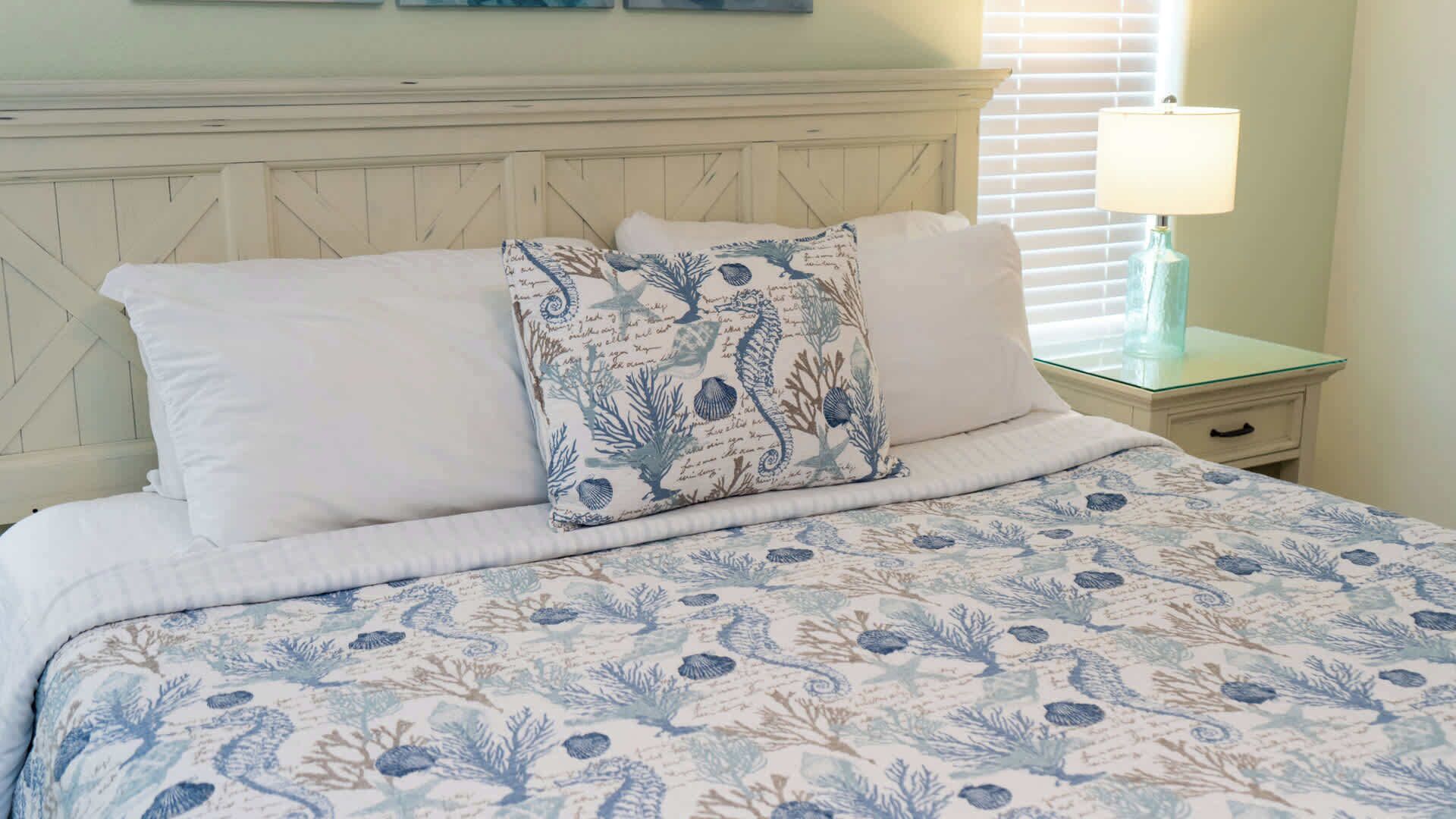 A bedroom with a bed, two pillows, a nightstand, a lamp, and a window with blinds.