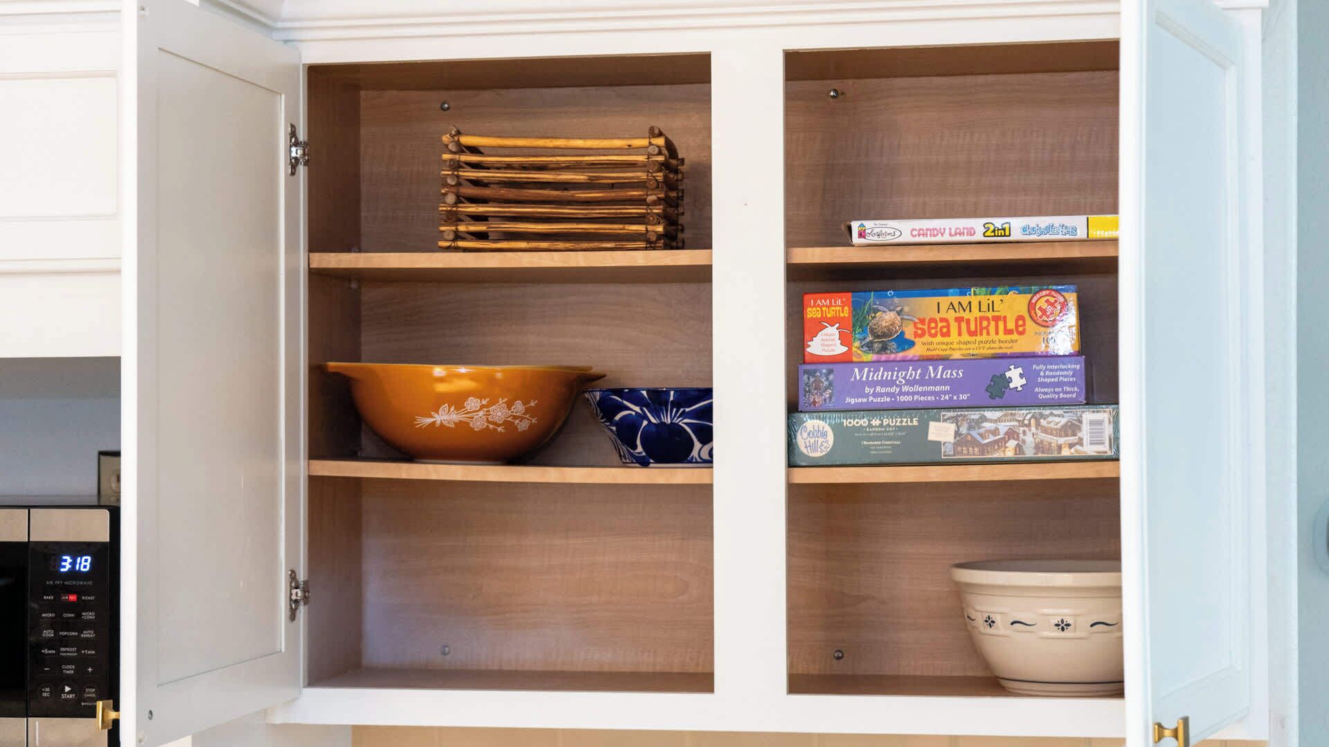 A kitchen cabinet with wooden shelves, a microwave, and various kitchenware including a bowl, a plate, and a box of puzzle pieces.