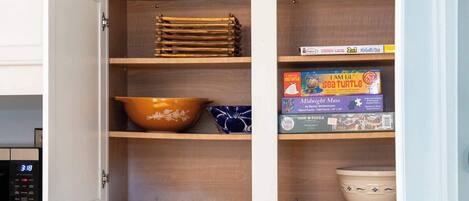 A kitchen cabinet with wooden shelves, a microwave, and various kitchenware including a bowl, a plate, and a box of puzzle pieces.