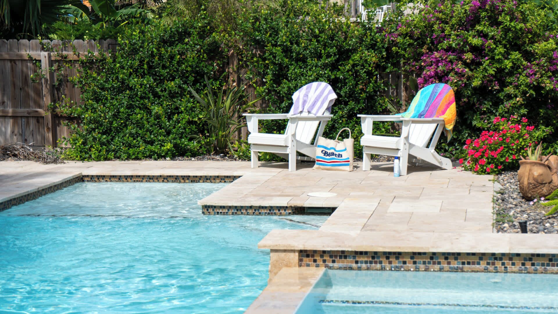 A swimming pool with two white lounge chairs, one with a striped towel and a bag, and the other with a colorful towel.