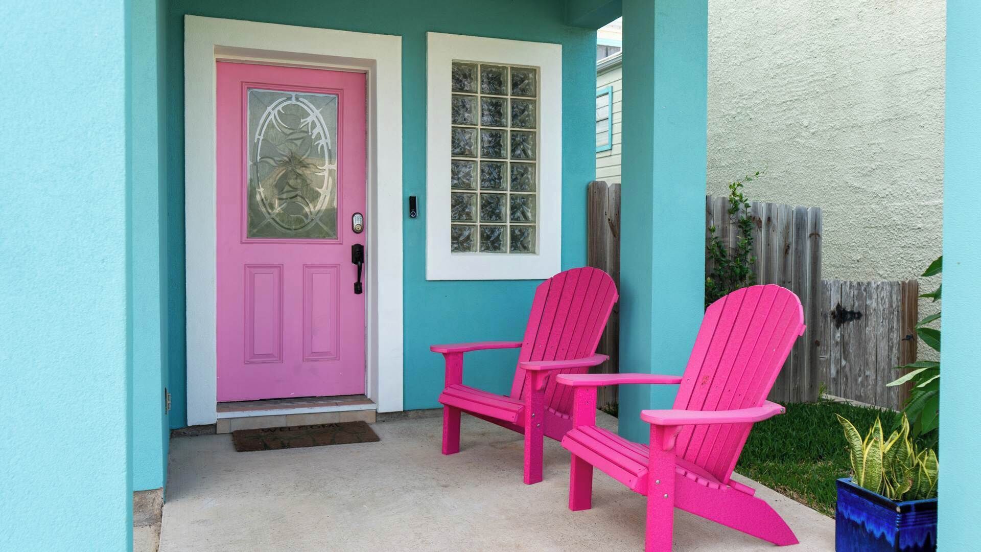 A turquoise house with a pink door and two pink Adirondack chairs outside.
