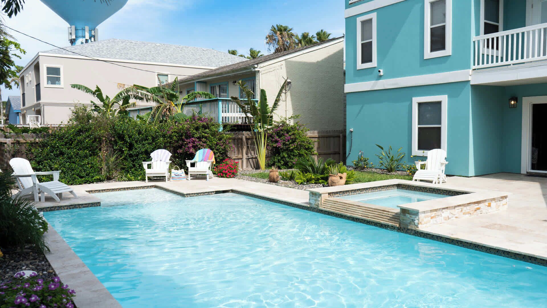 A swimming pool with two sections, surrounded by a garden with various plants and flowers. There are white lounge chairs and a blue building in the background.