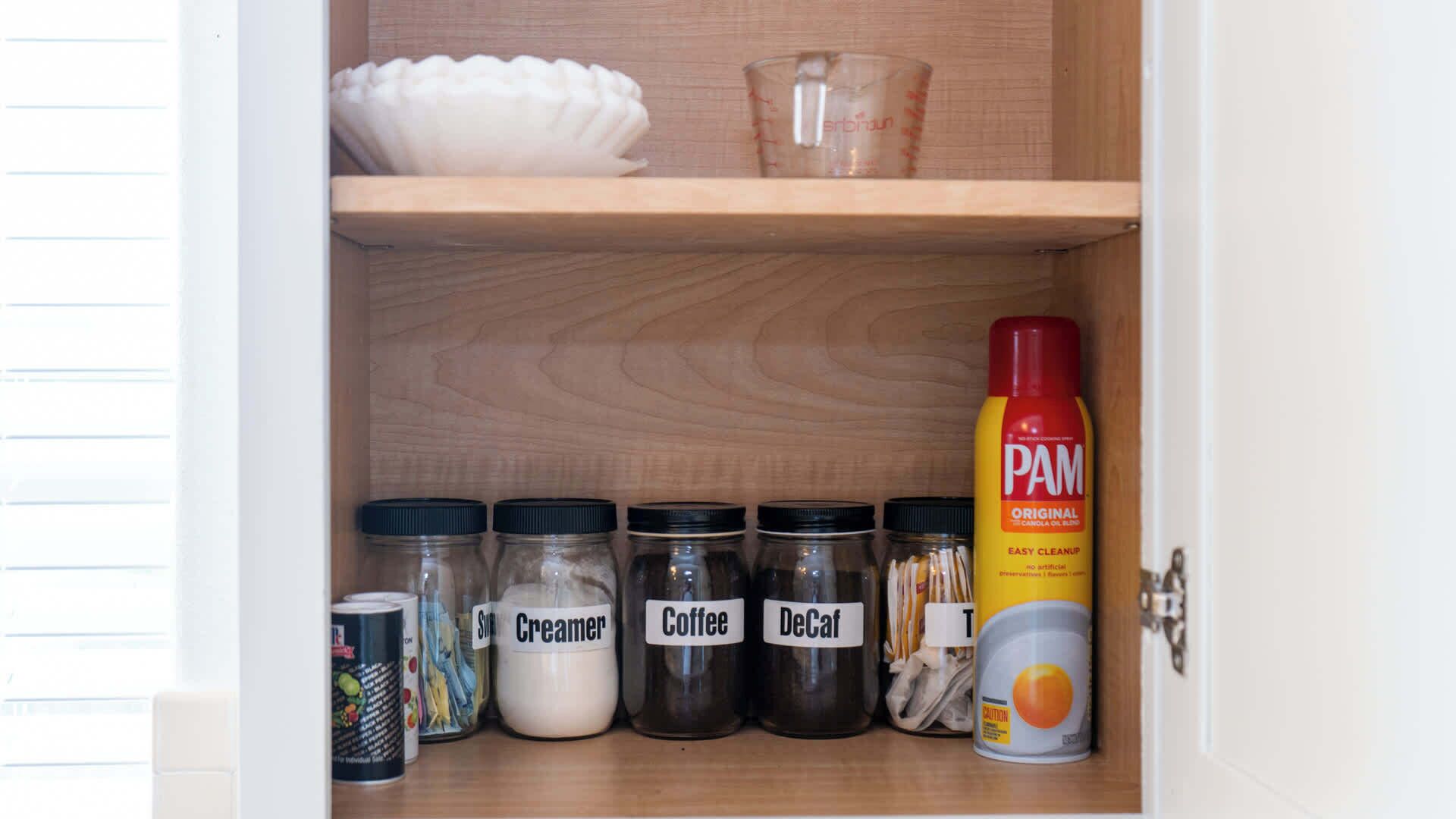A kitchen cabinet with jars labeled Creamer, Coffee, and DeCal, a bottle of PAM Original, and a can of coffee.