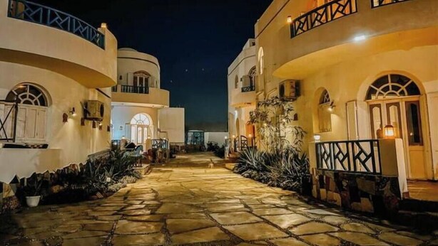 Una vista nocturna de un patio con senderos de piedra, edificios blancos con ventanas arqueadas y un cielo estrellado.