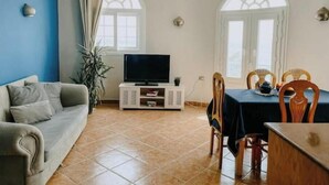 Una sala de estar con sofá, televisor, mesa de comedor con sillas y dos ventanas con cortinas.