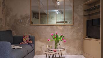 A living room with a sofa, a small round table with a vase of flowers, a mirror, and a television on a cabinet.