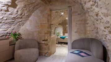 A cozy room with a stone wall, a bed, a chair, and a small table with a vase of flowers.