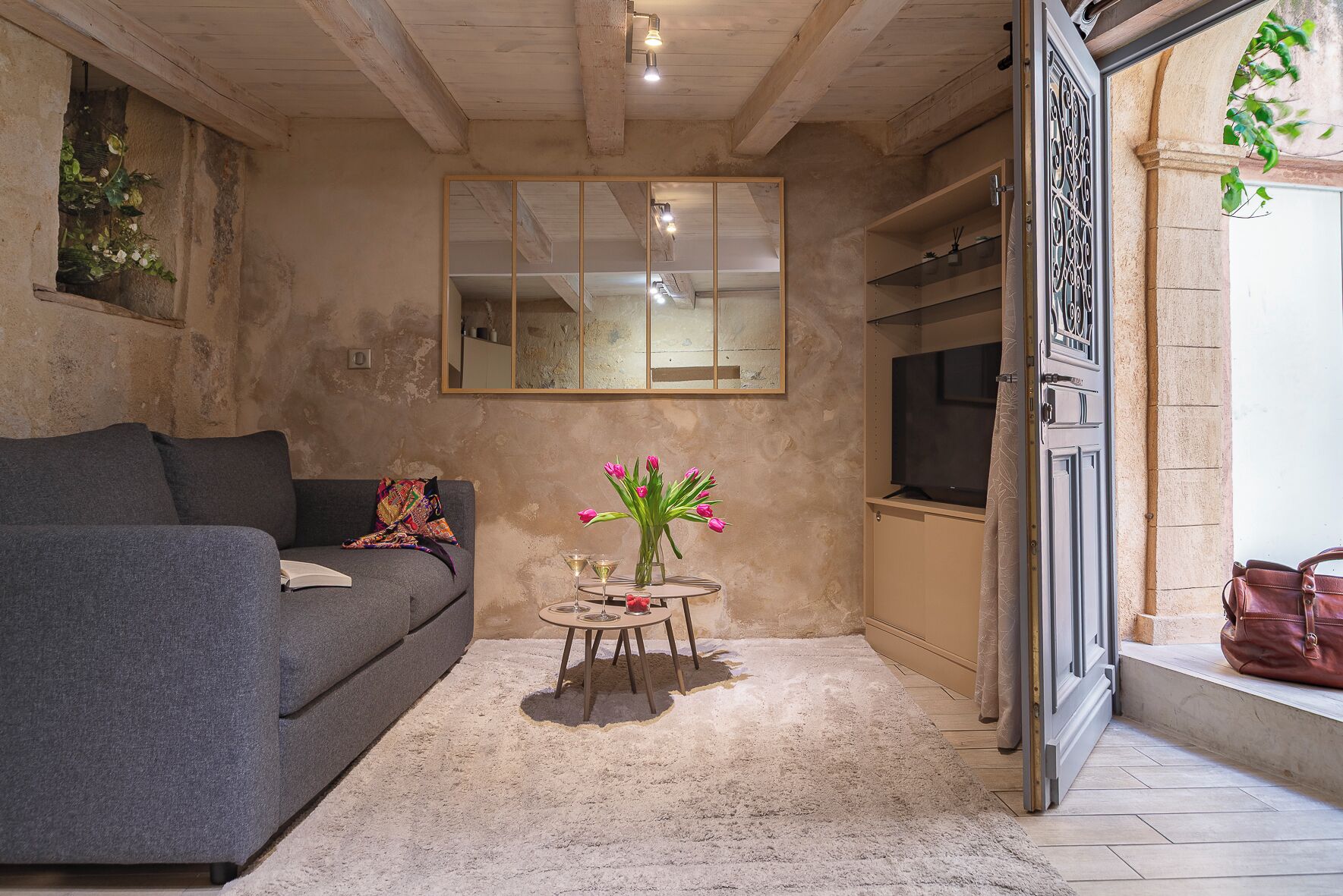 Ein Wohnzimmer mit einem grauen Sofa, einem kleinen runden Tisch mit Getränken, einem Spiegel und einem Fernseher auf einem Schrank.
