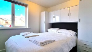 A neatly made bed with white linens and towels, a window with a view of buildings, and a wardrobe with sliding doors. - Beachfront apartment with direct beach access! (Perranporth)