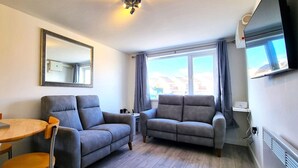 A modern living room with a grey sofa, a wooden dining table, and a large window with curtains. - Beachfront apartment with direct beach access! (Perranporth)