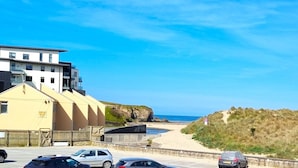Un bâtiment situé sur la côte, doté d’un toit plat, un parking avec plusieurs voitures et une plage de sable avec une falaise en arrière-plan.