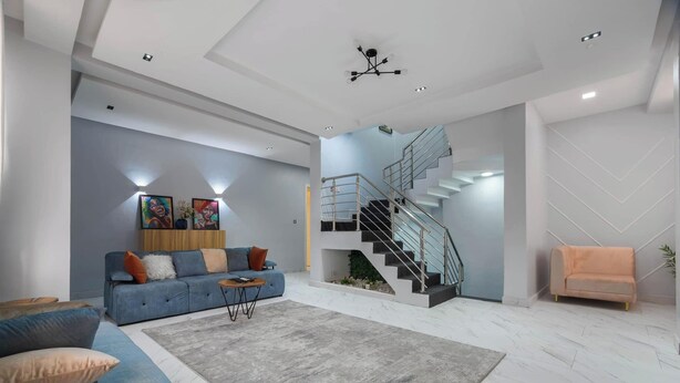 A modern living room with a blue sofa, a grey rug, a staircase, and a small orange armchair. - Blue Bell Suites and Apartments (Lekki)