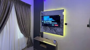 A modern living room with a flat-screen TV mounted on the wall, a dark entertainment unit below, and a large window with curtains. - Blue Bell Suites and Apartments (Lekki)