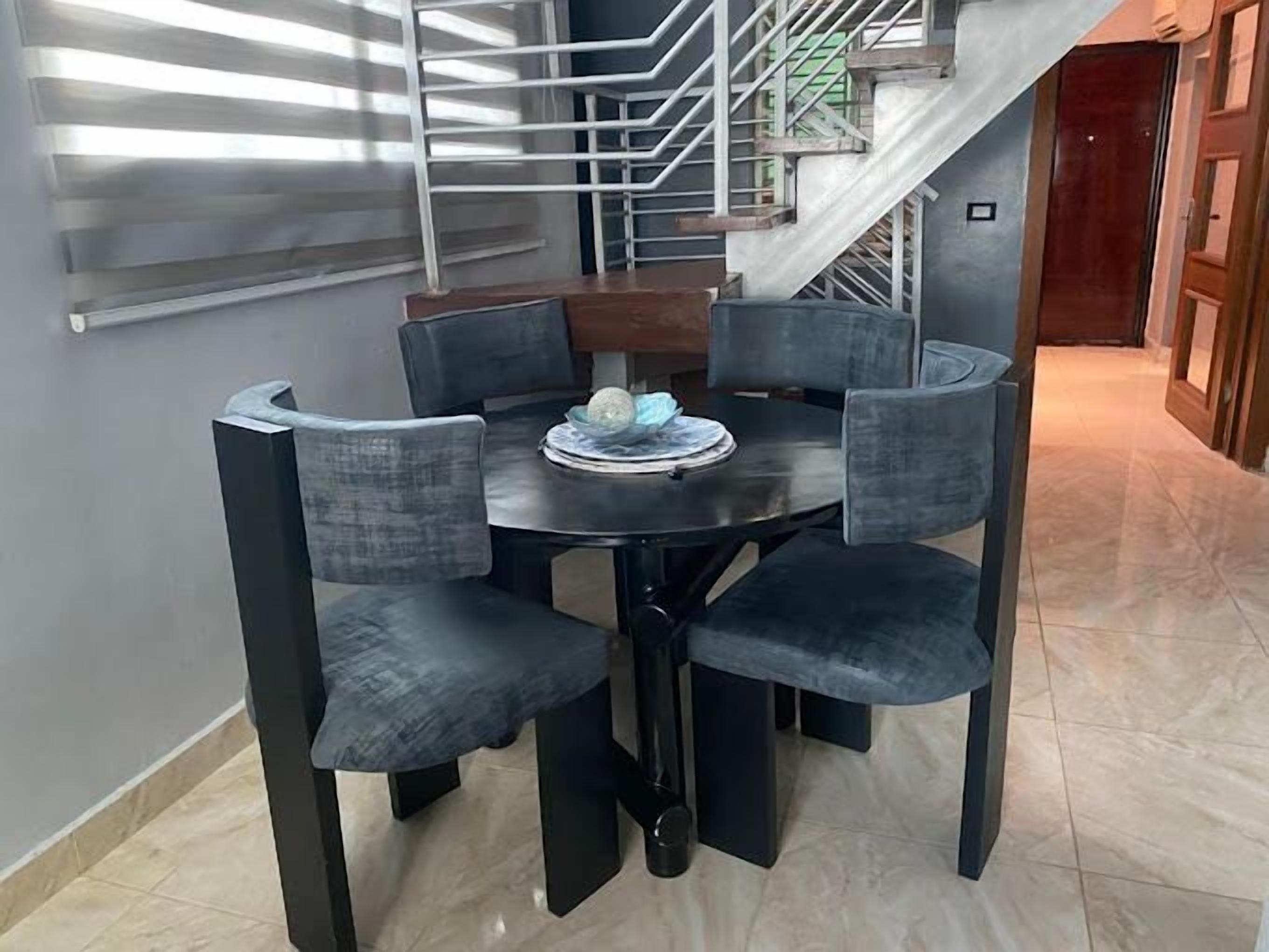 Un coin repas avec une table ronde et six chaises, un escalier en arrière-plan et une vue sur une porte en bois.