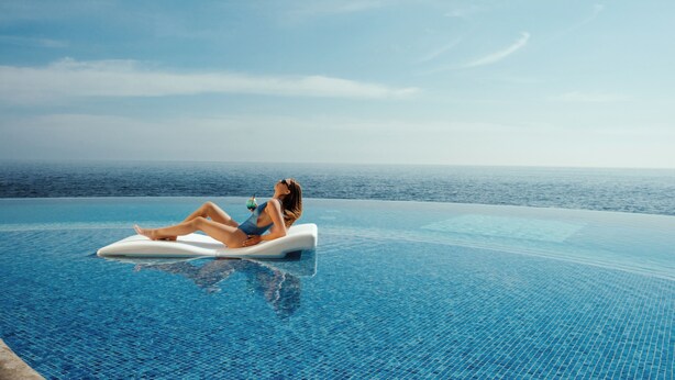 Una persona se relaja en una plataforma flotante en una piscina infinita de color azul intenso, con vistas al océano.