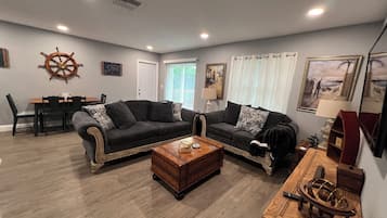 A living room with two sofas, a coffee table, a dining table with chairs, a ship's wheel decoration, and a painting on the wall.