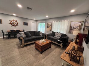 A living room with two sofas, a coffee table, a dining table with chairs, a ship's wheel decoration, and a painting on the wall.