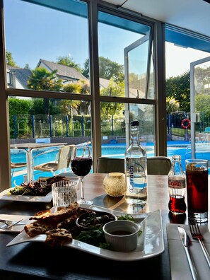 A dining table with a view of a swimming pool, a bottle of sparkling water, a glass of red wine, and a glass of Coca-Cola. - Haven (pet friendly) - 5 star self catering cottage (Carnon Downs)