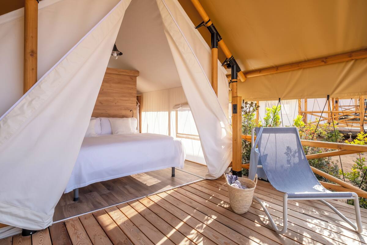 A wooden deck with a bed under a canopy, a chair, and a view of greenery.