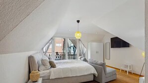 A modern loft bedroom with a sloped ceiling, a large bed, a grey sofa, and a view of a balcony. - Cozy half-timbered apartment in the center of Lüdinghausen with balcony (Lüdinghausen)