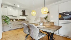 A modern kitchen with white cabinets, a wooden dining table, and two grey chairs. There are hanging lights and a plant on the counter. - Cozy half-timbered apartment in the center of Lüdinghausen with balcony (Lüdinghausen)