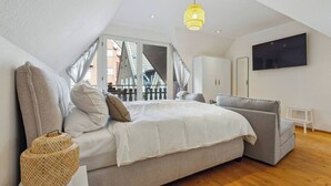 A bedroom with a bed, a grey sofa, a TV, and a window with a view of buildings. - Cozy half-timbered apartment in the center of Lüdinghausen with balcony (Lüdinghausen)