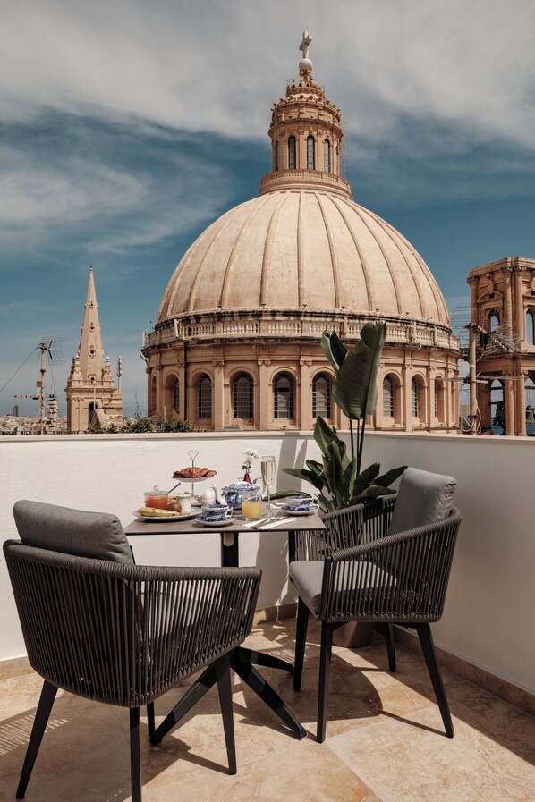 A terrace with two black wicker chairs, a table with breakfast items, and a view of a large dome building. - 1926 La Galerie Boutique Art Suites (Valletta)