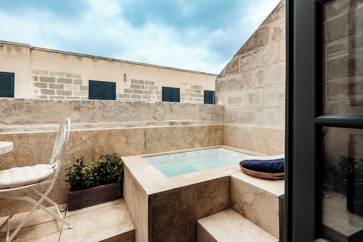 Un espacio al aire libre con paredes de piedra, una pequeña piscina, una silla acolchada y una planta en maceta.