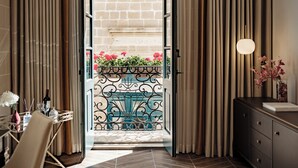 Puerta abierta que da a un balcón con reja de hierro forjado, un área de comedor con mesa y sillas, y un aparador con un florero.