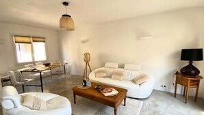 Ein modernes Wohnzimmer mit einem weißen Sofa, einem weißen Sessel, einem Holztisch und einem Essbereich mit einem Glastisch und Stühlen.
