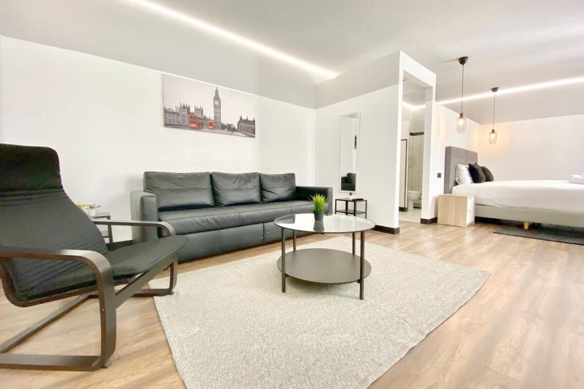 A modern living room with a grey sofa, a round coffee table, and a bed in the adjacent room.
