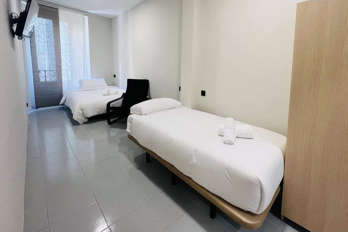 A hotel room with two single beds, a chair, a wooden wardrobe, and a window with a view.