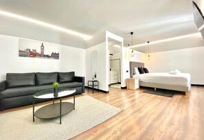 Un hotel moderno con un sofá de cuero negro, una mesa de centro redonda, una cama blanca y un cuadro de paisaje urbano en la pared.
