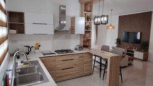 A modern kitchen with a central island, built-in cabinets, a stainless steel range hood, and a flat-screen TV mounted on a wooden panel wall.