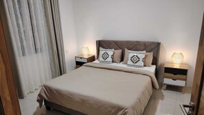 A neatly made bed with a neutral-colored bedspread and matching pillows, flanked by two bedside tables with lamps, in a room with a plain white wall and a partially visible curtain.