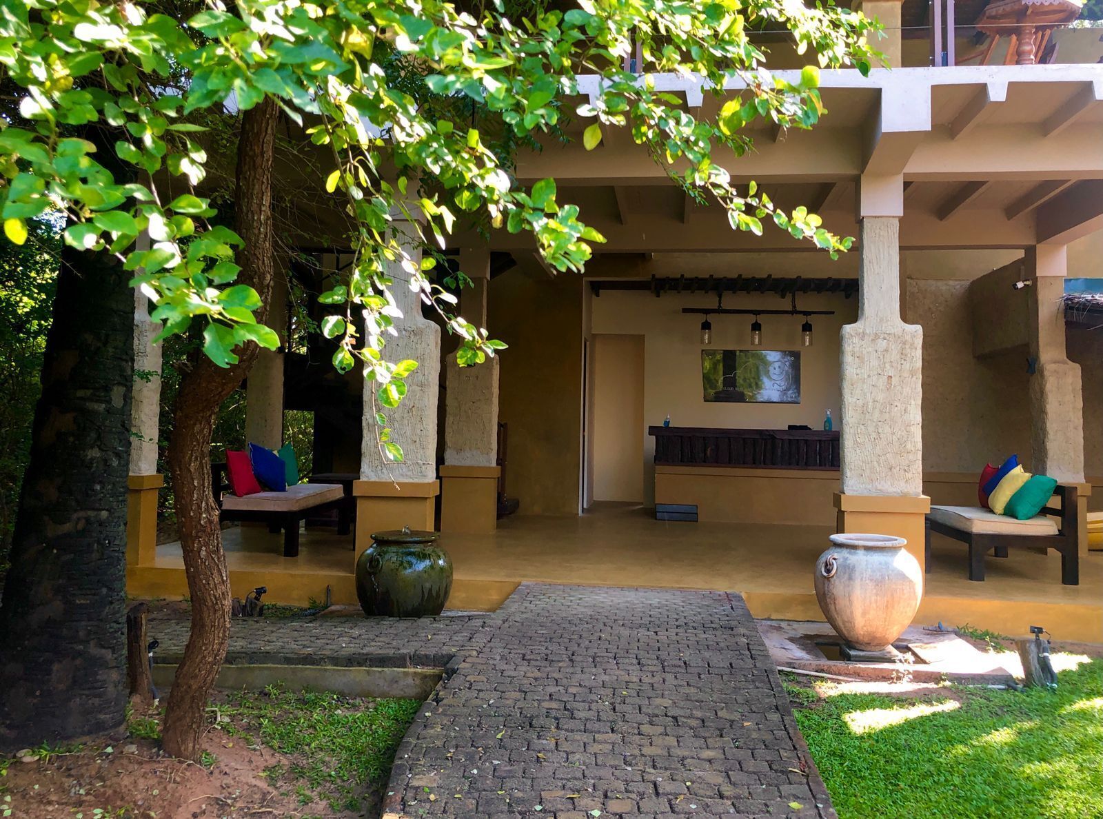 A covered outdoor area with a brick pathway leading to a seating area under a tree.