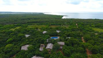 An aerial view of a tropical resort with multiple thatched-roof cottages, a central swimming pool, and a backdrop of dense forest and a large body of water.