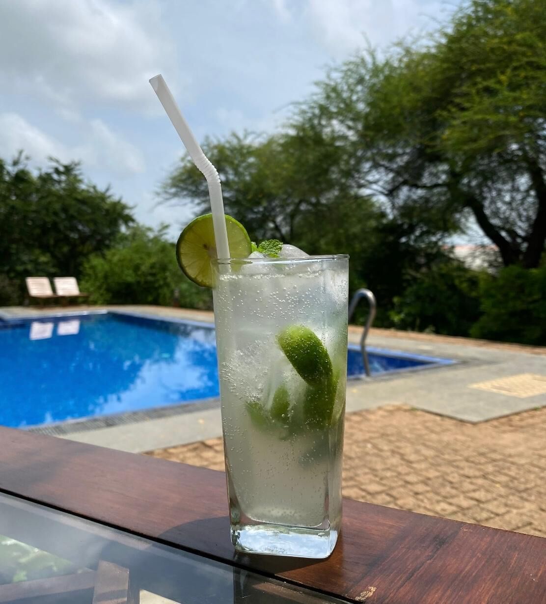 Un vaso de bebida helada con un gajo de lima y hojas de menta, sobre una mesa de madera cerca de una piscina.
